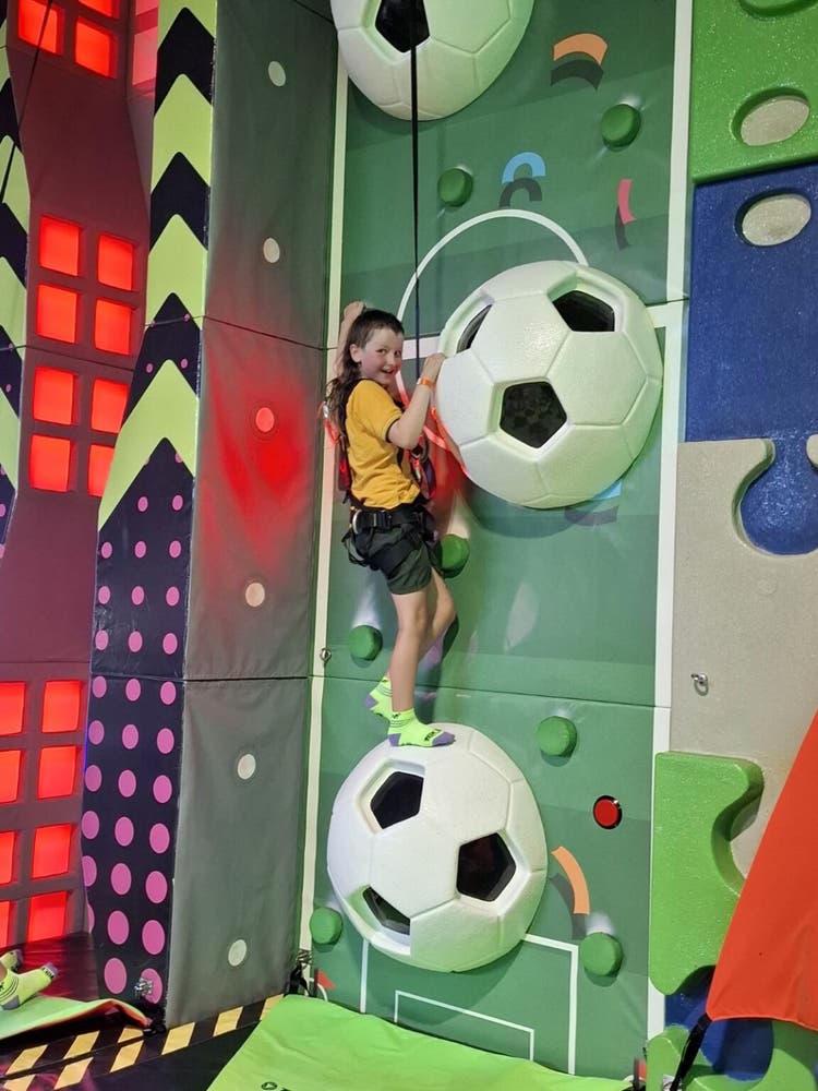 A male student climbing up a very colourful rock wall. He is looking at the camera, smiling while holding onto a shape coming out of the wall.