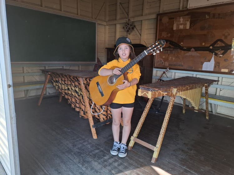 student holding a guitar in the music shed. In the background there are marimbas and a piano